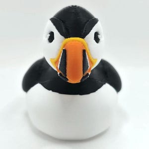 May include: A 3D printed puffin figurine featuring a black and white body with a prominent, multi-colored beak in shades of orange, yellow, and black. The bird has a rounded, smooth shape with visible horizontal layer lines from the printing process.