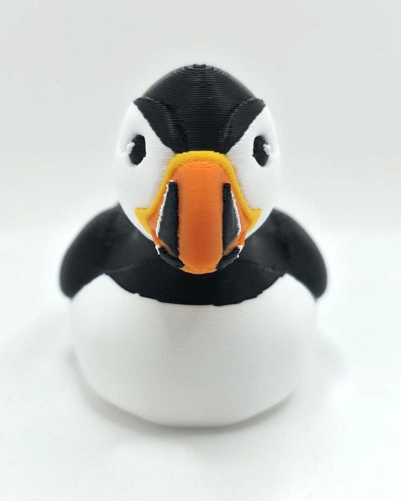 May include: A 3D printed puffin figurine featuring a black and white body with a prominent, multi-colored beak in shades of orange, yellow, and black. The bird has a rounded, smooth shape with visible horizontal layer lines from the printing process.