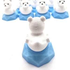 May include: A collection of small, 3D-printed white bear figurines, each perched atop a light blue hexagonal base. The bears feature rounded ears and simple facial details, with one bear positioned in the foreground facing away from the viewer.