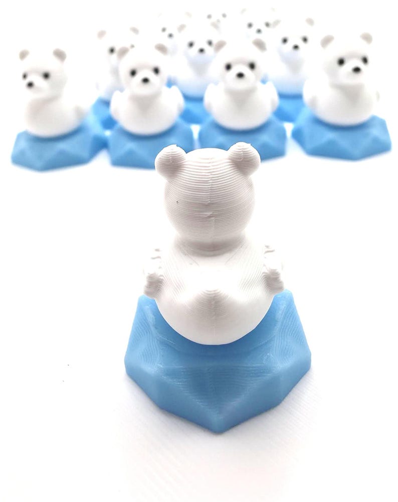 May include: A collection of small, 3D-printed white bear figurines, each perched atop a light blue hexagonal base. The bears feature rounded ears and simple facial details, with one bear positioned in the foreground facing away from the viewer.