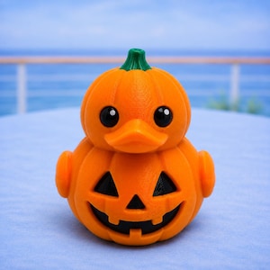 May include: A bright orange rubber duck shaped like a pumpkin with a green stem on top. The duck has large black eyes and a carved jack-o'-lantern face on its belly with triangular eyes and a jagged mouth showing a dark interior cavity.