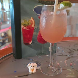 May include: A stemmed glass filled with a pale pink cocktail, ice cubes, and a white straw, garnished with a lime wedge. Beside the glass sits a small white elephant figurine. A menu featuring images of colorful cocktails stands in the background.