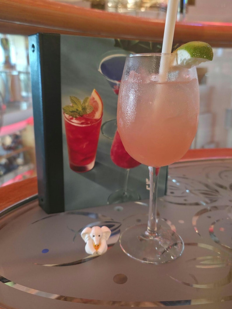 May include: A stemmed glass filled with a pale pink cocktail, ice cubes, and a white straw, garnished with a lime wedge. Beside the glass sits a small white elephant figurine. A menu featuring images of colorful cocktails stands in the background.