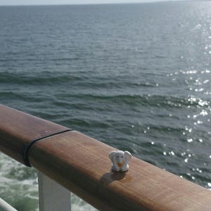 May include: A small white elephant figurine with a yellow accent on its trunk, positioned on a smooth, polished wooden railing. The surface of the railing shows a natural wood grain texture, and the object is captured in bright daylight.