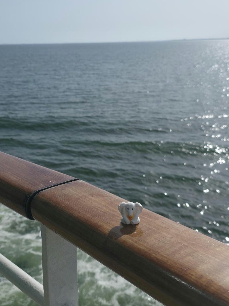 May include: A small white elephant figurine with a yellow accent on its trunk, positioned on a smooth, polished wooden railing. The surface of the railing shows a natural wood grain texture, and the object is captured in bright daylight.