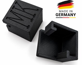 Könnte beinhalten: Zwei schwarze, 3D-gedruckte quadratische Halterungen mit strukturierter Oberfläche. Eine Halterung steht aufrecht und zeigt die geprägten Buchstaben MK auf der Oberseite. Die andere Halterung liegt auf der Seite und zeigt das offene, hohle Innere.