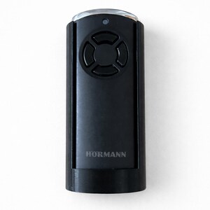 Könnte beinhalten: Schwarze Hormann Garagentor-Fernbedienung. Die Fernbedienung hat eine rechteckige Form mit abgerundeten Kanten und einem kreisförmigen Tastenlayout. Der Markenname "HORMANN" ist auf der Vorderseite aufgedruckt.