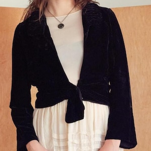 May include: A black velvet jacket with long sleeves and a tie closure. The jacket has a shawl collar and is worn over a cream-colored dress. The overall style is vintage and elegant.