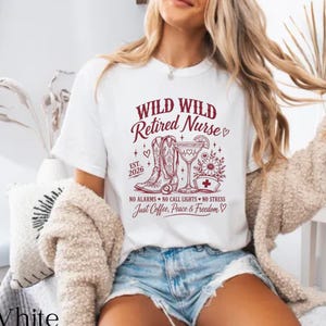 May include: A white short-sleeved t-shirt featuring a maroon graphic print with the text Wild Wild Retired Nurse, Est. 2026, No Alarms, No Call Lights, No Stress, and Just Coffee, Peace & Freedom, alongside illustrations of cowboy boots and a drink.