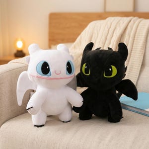 May include: Two plush dragon toys, one white with light blue eyes and a pink mouth line, and one black with bright green eyes. Both feature large heads, pointed ears, small wings, and rounded bodies, crafted from soft fabric materials.