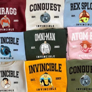 Puede incluir: Una colecci&oacute;n de sudaderas de cuello redondo en varios colores, cada una con un personaje diferente de la serie de c&oacute;mics Invincible. Las sudaderas muestran nombres de personajes, la palabra "Invincible" y el a&ntilde;o "2003". Cada dise&ntilde;o incluye una ilustraci&oacute;n circular del personaje.