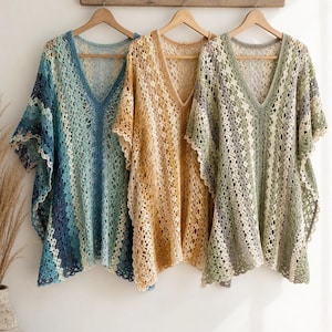 May include: Three open-weave, V-neck tops in shades of blue, gold, and green hang on a wooden rack. The tops have a loose, flowing design with a scalloped edge. The crochet pattern creates a delicate, airy look.