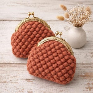 Könnte beinhalten: Zwei terrakottafarbene Häkeltaschen mit goldfarbenen Verschlüssen. Die Taschen haben ein strukturiertes Noppenmuster. Eine Tasche befindet sich vor der anderen. Im Hintergrund eine kleine weiße Vase mit Trockenblumen.