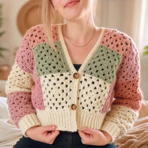 May include: A handmade crochet cardigan in a patchwork design. The sweater features a V-neck and is buttoned up. The color scheme includes cream, dusty rose, and sage green squares. The sleeves are long and the cardigan is made of soft yarn.