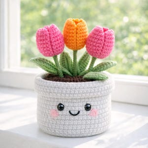 Puede incluir: Una maceta blanca tejida con una cara sonriente contiene tres tulipanes coloridos. Los tulipanes son rosas y naranjas, con tallos y hojas verdes. La maceta está sobre una superficie blanca, con una ventana en el fondo.