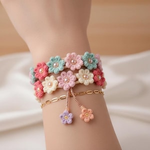 Könnte beinhalten: Ein Blumenarmband mit gehäkelten Blumen in Rosa, Creme und Blau. Das Armband hat eine goldene Kette mit baumelnden Blumenanhängern. Die Blumen sind mit kleinen perlenartigen Perlen verziert.