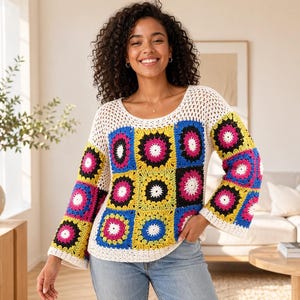 Può includere: Un colorato maglione all'uncinetto con un design patchwork. Il maglione presenta uno sprone e maniche a trama aperta bianche, con pannelli quadrati gialli, rosa, blu e neri. La modella indossa jeans blu.