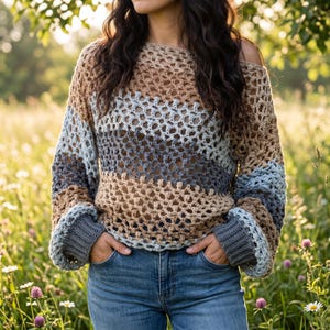Peut inclure: Un pull en crochet à manches longues présentant un motif à mailles ouvertes et lâches avec des rayures horizontales dans des tons de beige, bleu clair et gris foncé. Le vêtement possède une large encolure bateau et des poignets côtelés gris foncé.