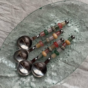 Könnte beinhalten: Vier Edelstahl-Löffel mit bunten Perlenstielen liegen auf einem ovalen, transparenten Glasteller. Die Perlen sind in den Farbtönen Rosa, Grün, Gelb und gesprenkeltem Beige gehalten. Jeder Löffel hat eine kleine, runde Schale und eine silberne Kugel am Ende des Stiels.