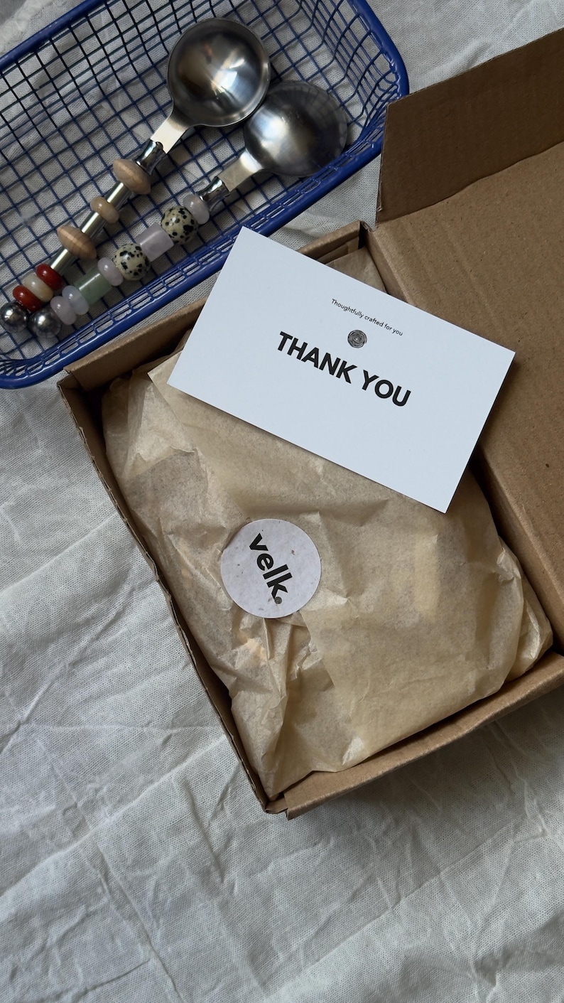 Pu&ograve; includere: Un cestino blu in filo metallico contiene due cucchiai da portata in acciaio inossidabile con manici decorativi. Una scatola di cartone contiene una carta con la scritta "THANK YOU" e un adesivo con la parola "velk."