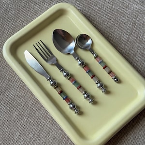 May include: A set of four stainless steel utensils including a knife, fork, large spoon, and small spoon, each featuring handles decorated with colorful stacked beads and silver-toned metal accents, all arranged on a pale yellow rectangular tray.