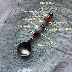 Pu&ograve; includere: Un cucchiaio decorativo con un manico ornato da varie pietre preziose, tra cui agata rossa, avventurina verde, quarzo rosa, tormalina, diaspro dalmata e sodalite. Il cucchiaio &egrave; appoggiato su un piatto di vetro.