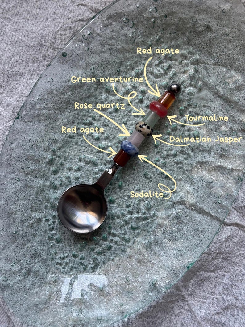 Pu&ograve; includere: Un cucchiaio decorativo con un manico ornato da varie pietre preziose, tra cui agata rossa, avventurina verde, quarzo rosa, tormalina, diaspro dalmata e sodalite. Il cucchiaio &egrave; appoggiato su un piatto di vetro.