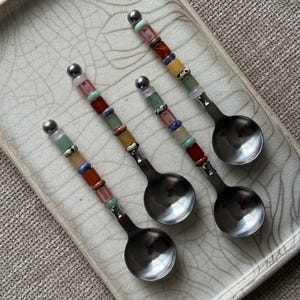 May include: Three silver-toned metal spoons with handles decorated with a series of multi-colored, cylindrical stone beads in shades of pink, green, blue, orange, and white, topped with a small silver-toned metal sphere at the end of each handle.