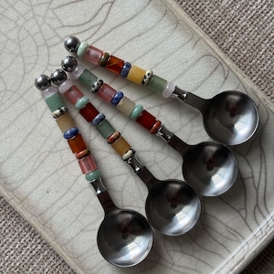 May include: Three metal spoons with handles decorated with a series of colorful, multi-shaped gemstone beads. The beads include shades of red, orange, yellow, green, blue, and spotted patterns, finished with a small round metal bead at the top.