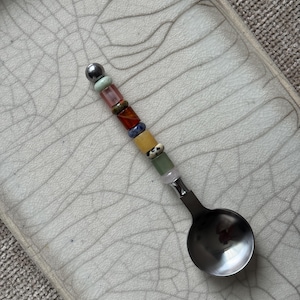 May include: A metal spoon featuring a handle decorated with a vertical row of multi-colored beads in shades of green, pink, brown, blue, yellow, and white, topped with a single metallic sphere, resting on a light-colored tray with a crackle glaze.