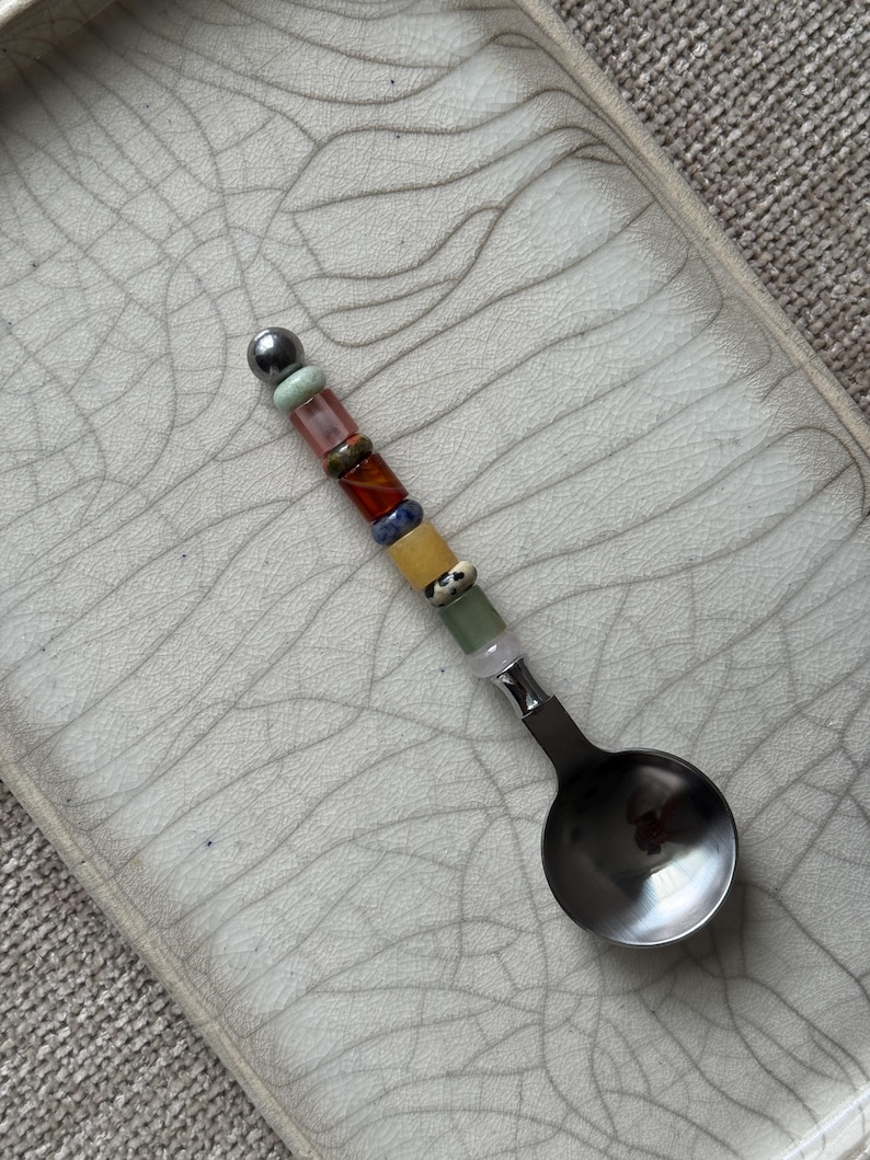 May include: A metal spoon featuring a handle decorated with a vertical row of multi-colored beads in shades of green, pink, brown, blue, yellow, and white, topped with a single metallic sphere, resting on a light-colored tray with a crackle glaze.