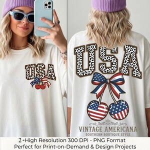 Op de afbeelding: Een wit t-shirt met een grafisch ontwerp met de tekst USA in een zwart-wit luipaardprint. Onder de tekst bevinden zich twee kersen met een Amerikaans vlagpatroon, samengebonden met een rood, wit en blauw gestreepte strik.