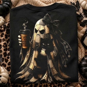 May include: A black t-shirt featuring a graphic of a ghost wearing a large bow and holding a coffee cup with the text 'WE'RE NOT MORNING PEOPLE'. A black cat is nestled in the ghost's arms. The design uses shades of gold, tan, and black.