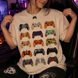Op de afbeelding: Een beige T-shirt met ronde hals voorzien van een raster met diverse illustraties van videogamecontrollers met Halloween-thema's, waaronder skeletten, pompoenen, zombies en vleermuizen, bedrukt in oranje, zwart, groen, paars en wit.
