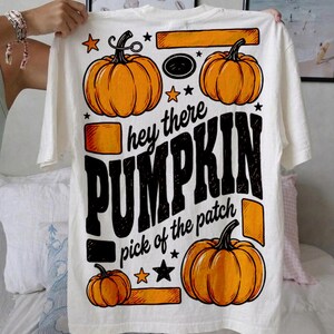 Puede incluir: Una camiseta blanca con un estampado gráfico de tres calabazas naranjas, estrellas negras y detalles rectangulares naranjas. El texto dice "hey there PUMPKIN" en letras negras, seguido de "pick of the patch" escrito debajo.