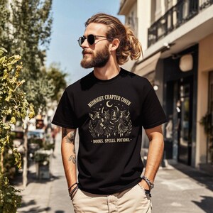 Op de afbeelding: Zwart T-shirt met de tekst "MIDNIGHT CHAPTER COVEN" boven een afbeelding van drie dansende figuren onder een halve maan. De tekst "BOOKS. SPELLS. POTIONS." staat eronder. Het shirt heeft korte mouwen.