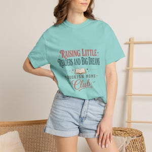 Pode incluir: Uma camiseta azul clara com o texto "Raising Little Readers and Big Dreams" e "Bookish Moms Club" em vermelho e azul. A camiseta é combinada com shorts jeans.