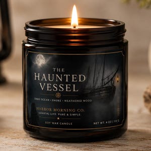 May include: A lit soy wax candle in a dark amber glass jar with a black label featuring a ship illustration and the text The Haunted Vessel, Harbor Morning Co., Dark Ocean, Smoke, Weathered Wood, Coastal Life, Pure & Simple, and Net Wt. 4 oz.
