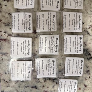May include: A collection of thirteen clear plastic clamshell packages containing wax melts from Harbor Morning Co. Each label displays a unique scent name, such as Yacht Club, Coastal Resort, and Strawberry Fields, along with ordering information.