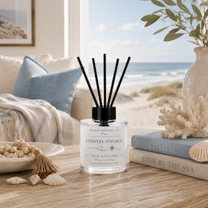 Op de afbeelding: Een glazen rietdiffuser met vijf zwarte rietjes. Het witte etiket vermeldt Harbor Morning Co. Coastal Voyage Reed Diffuser 100 ml / 3.4 fl oz. De fles staat op een houten ondergrond naast een schaal met houten kralen en twee boeken.