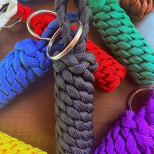 May include: A collection of braided rope keychains in various colors, including black, blue, red, green, yellow, purple, white, and brown. Each keychain features a silver metal ring.