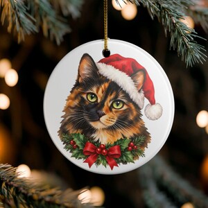 May include: A round white ceramic ornament featuring a detailed illustration of a tortoiseshell cat wearing a red and white Santa hat. The cat is framed by a festive wreath of green holly leaves and red berries with a red bow at the bottom.