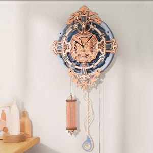 Puede incluir: Un reloj decorativo de madera con un diseño azul claro y color cobre. El reloj presenta números romanos, intrincados detalles recortados y un péndulo con un cilindro rosa y una forma de lágrima azul.