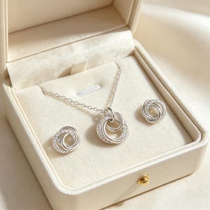 May include: A silver jewellery set comprising a necklace and stud earrings, displayed in a cream box. The necklace has a pendant with two interlocking circles, each set with small, clear crystals. The earrings echo the pendant's design, creating a coordinated look.
