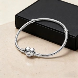 Op de afbeelding: Een zilveren armband met een hartvormige bedel met de inscriptie "PANDORA". De armband ligt op een licht oppervlak, met een zwarte doos op de achtergrond.