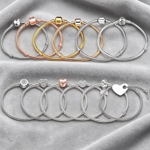 Könnte beinhalten: Eine Sammlung von Armbändern mit Charms in Silber, Gold und Roségold. Die Armbänder haben verschiedene Charms, darunter Herzen, Perlen und einen Charm mit dem Wort "love". Die Armbänder werden auf einer grauen Oberfläche präsentiert.