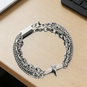 Może przedstawiać: Srebrna bransoletka z wieloma stylami łańcuchów. Bransoletka zawiera prostokątną płytkę i akcent w kształcie gwiazdy z centralnym klejnotem. Łańcuchy różnią się grubością i wzorem, tworząc warstwowy efekt.