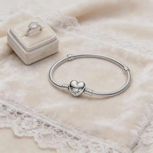 May include: A silver snake chain bracelet featuring a heart-shaped clasp with the engraved text PANDORA, accompanied by a silver ring with a round white stone set in a small square velvet ring box with a lid.