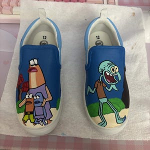 Op de afbeelding: Een paar blauwe instapschoenen met handgeschilderde figuren uit SpongeBob SquarePants. De linkerschoen toont een lange oranje vis en twee kleinere vissen, terwijl de rechterschoen een groene inktvisfiguur bevat. Beide inlegzolen zijn gemarkeerd met maat 12.