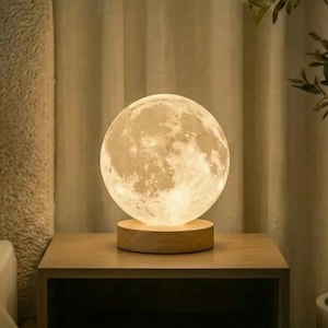 May include: A glowing moon-shaped lamp sits on a wooden base, placed on a nightstand. The lamp emits a soft, warm light, creating a cozy ambiance. The lamp's surface mimics the moon's craters and textures. The nightstand is made of light-colored wood.